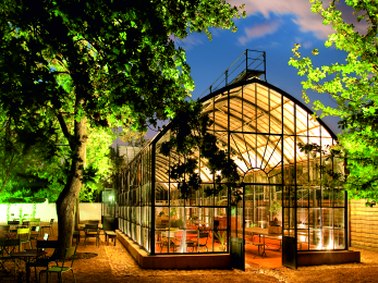 Iconic, night view of Babylonstoren’s famous glasshouse restaurant, glowing warmly with outdoor tables and chairs under mature trees