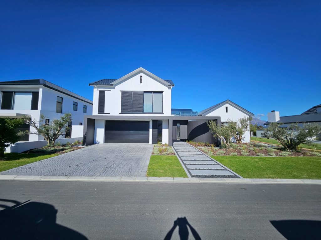 Street view of a contemporary home at The Acres, Pearl Valley, Paarl, with multi-stem olive trees, silver groundcover planting, lawn, grey stepping-stone path and herringbone driveway by CSA Landscaping, best landscapers in the Boland and Cape Winelands