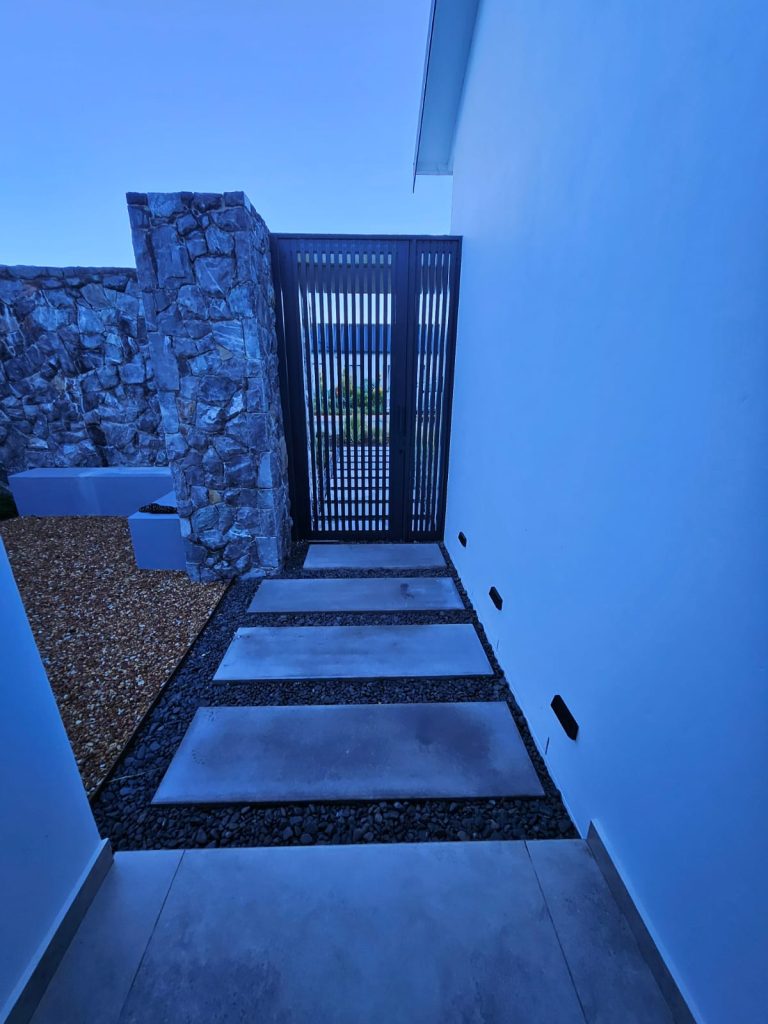 Entrance pathway with grey stepping stones, black metal gate and natural stone cladding by CSA Landscaping, the best in Boland and Cape Winelands landscaping, at The Acres at Pearl Valley, Paarl