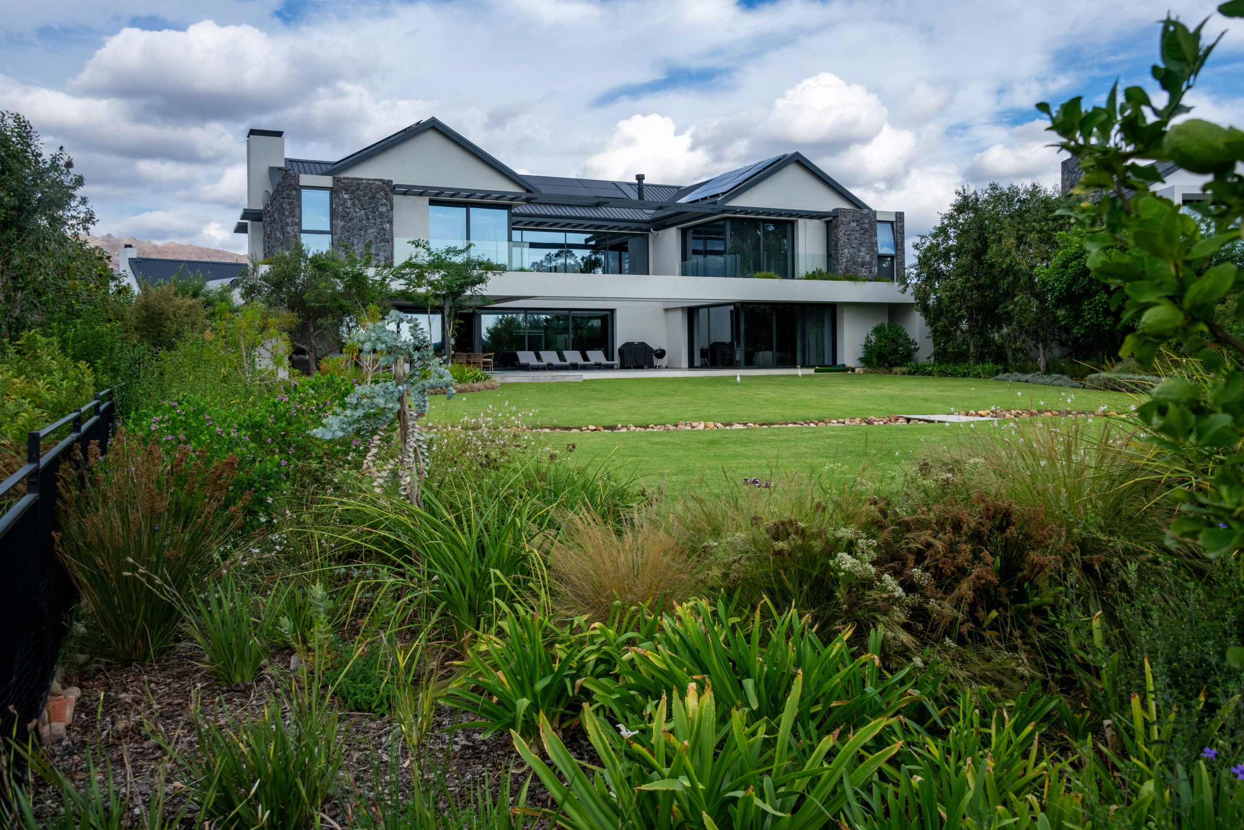 Foreground planting of Dietes, ornamental grasses and mixed shrubs with bark mulch, looking across the lawn to a contemporary home at Val de Vie Estate, Paarl, by CSA Landscaping — leading landscapers in the Boland and Cape Winelands