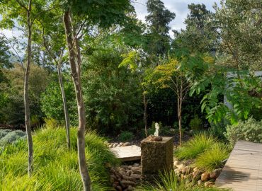 Granite water feature set in ornamental grasses and river stones, with timber boardwalk and feature trees at Val de Vie Estate, Paarl, by CSA Landscaping — leading landscapers in the Boland and Cape Winelands