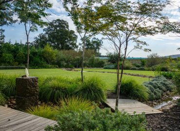 Granite water feature and timber boardwalk over the circulating stream, with ornamental grasses, Helichrysum and open lawn at Val de Vie Estate, Paarl, by CSA Landscaping — leading landscapers in the Boland and Cape Winelands