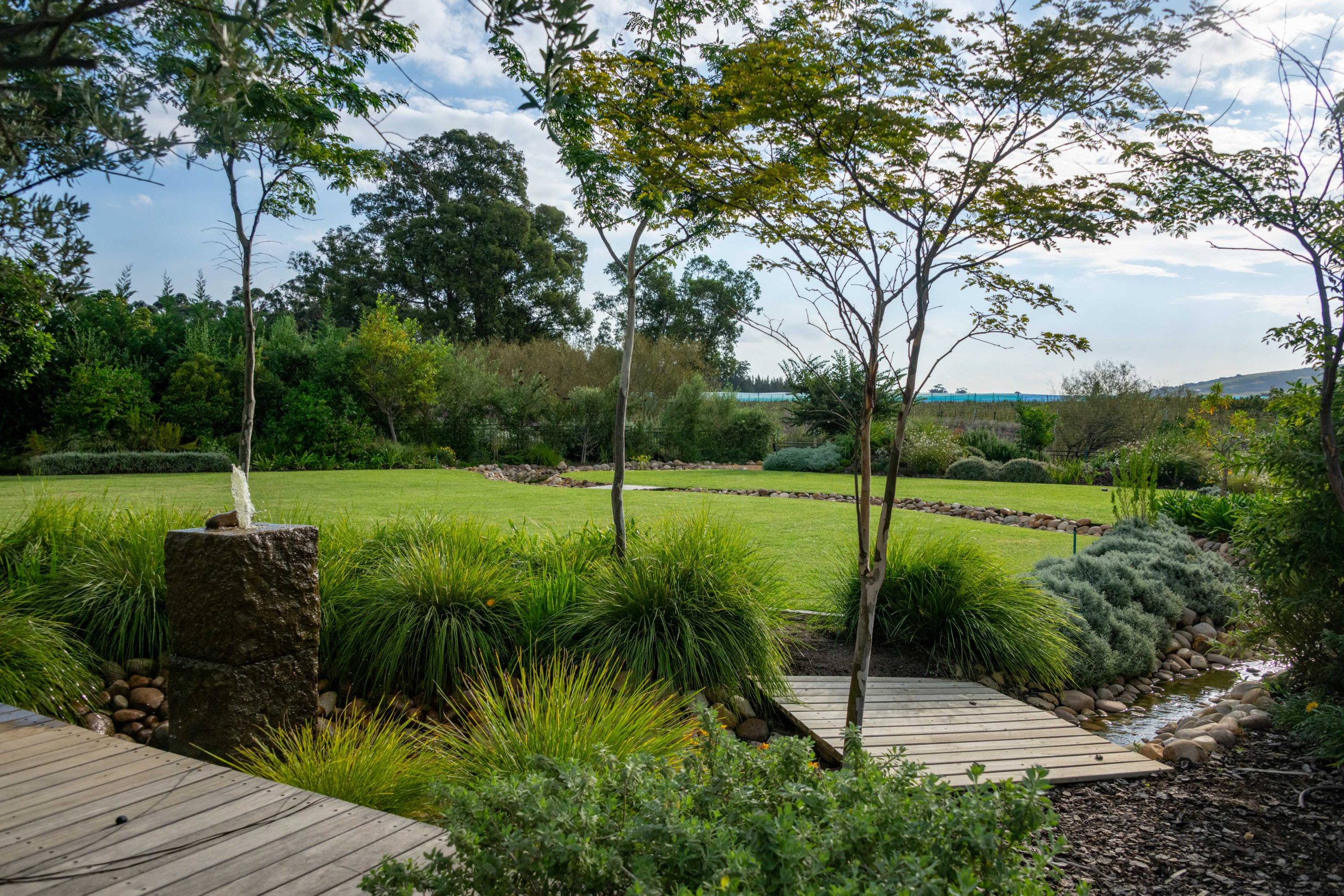 Granite water feature and timber boardwalk over the circulating stream, with ornamental grasses, Helichrysum and open lawn at Val de Vie Estate, Paarl, by CSA Landscaping — leading landscapers in the Boland and Cape Winelands