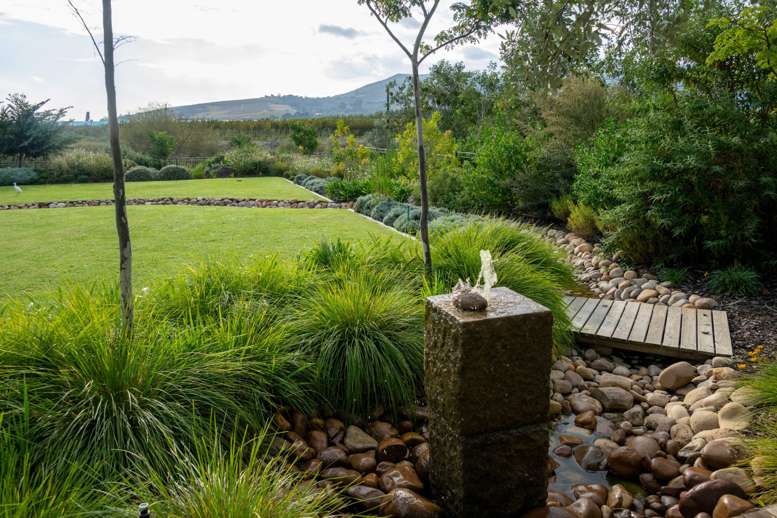 Custom granite water feature feeding the river-stone stream, with timber bridge, ornamental grasses and lawn at Val de Vie Estate, Paarl, by CSA Landscaping — leading landscapers in the Boland and Cape Winelands