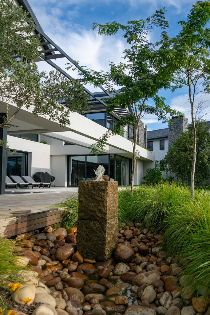Custom granite water feature by CSA Landscaping, with water flowing over river stones beside a timber boardwalk, olive trees and ornamental grasses at Val de Vie Estate, Paarl — leading landscapers in the Boland and Cape Winelands