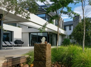 Custom granite water feature by CSA Landscaping, with water flowing over river stones beside a timber boardwalk, olive trees and ornamental grasses at Val de Vie Estate, Paarl — leading landscapers in the Boland and Cape Winelands