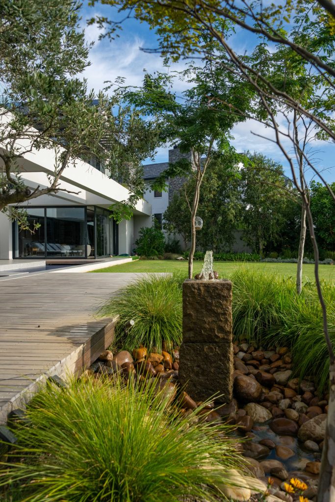 Custom granite water feature flowing onto river stones beside a timber boardwalk, framed by olive trees and ornamental grasses at Val de Vie Estate, Paarl, by CSA Landscaping — leading landscapers in the Boland and Cape Winelands