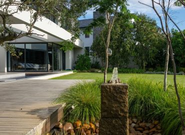 Custom granite water feature flowing onto river stones beside a timber boardwalk, framed by olive trees and ornamental grasses at Val de Vie Estate, Paarl, by CSA Landscaping — leading landscapers in the Boland and Cape Winelands