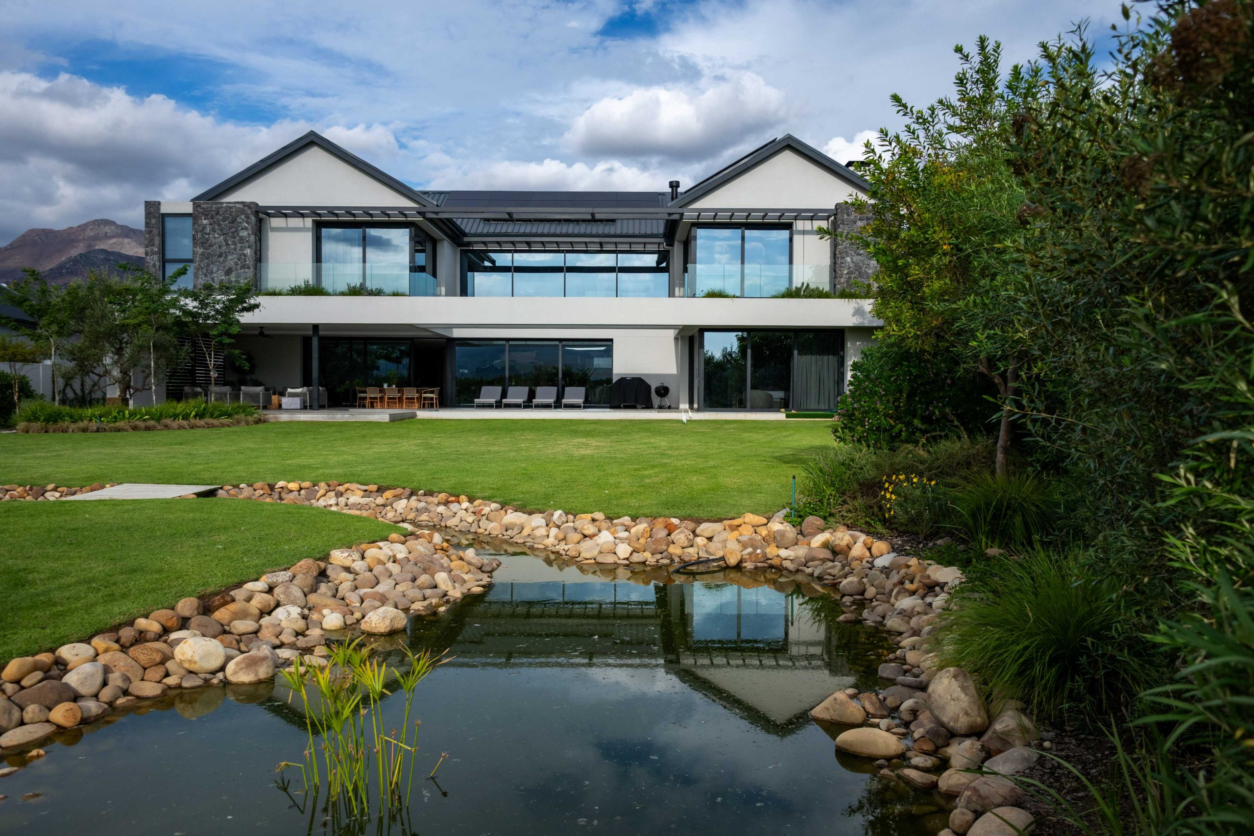 Lawn edged with silver Helichrysum and white-flowering Gaura, with Dietes and a river-stone stream leading to a contemporary home in Val de Vie Estate, Paarl, by CSA Landscaping — leading landscapers in the Boland and Cape Winelands